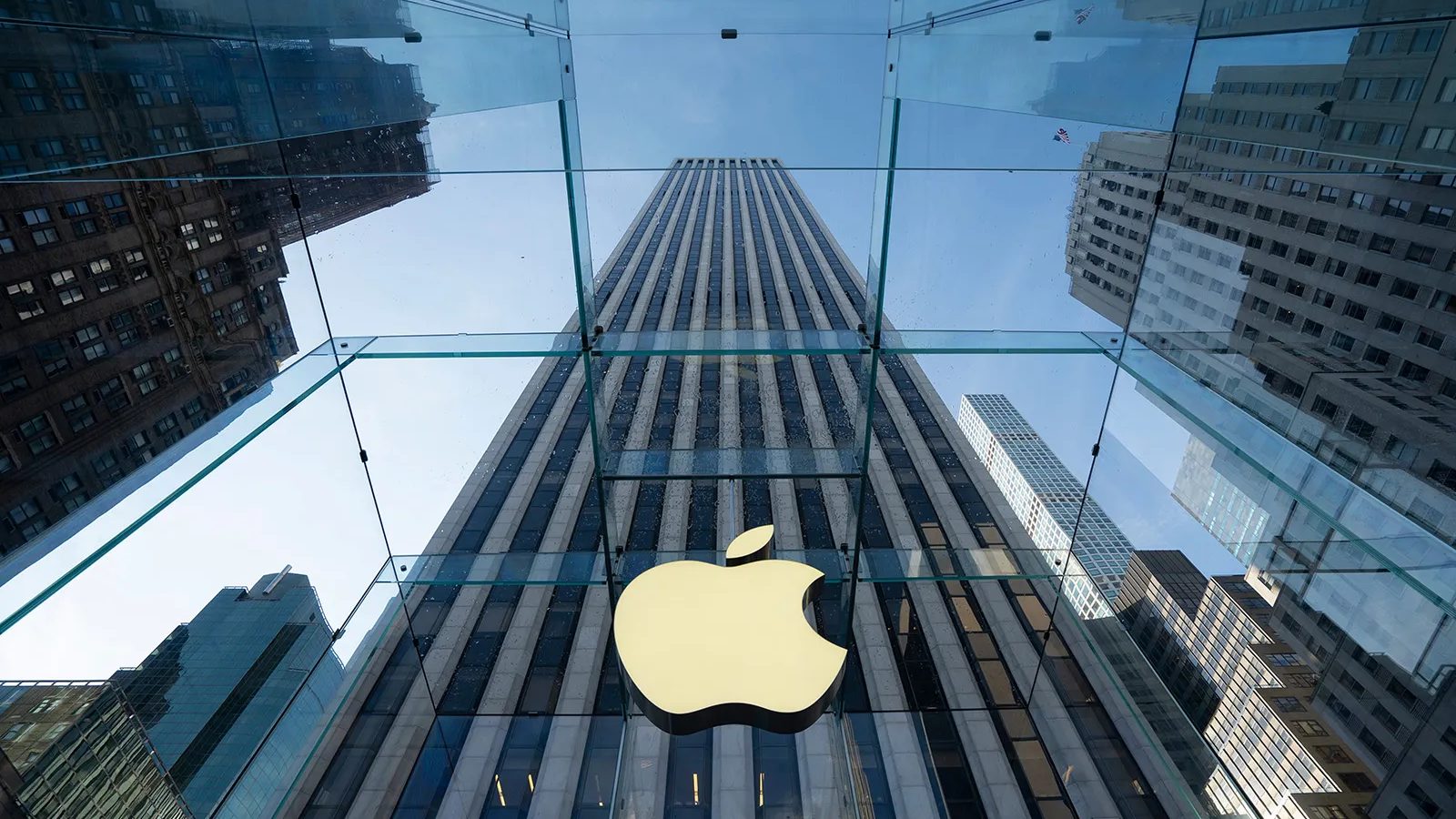 Looking up at the glass cube entrance of the Apple Store on Fifth Avenue in New York, with the illuminated Apple logo suspended inside and skyscrapers reflected in the panels.