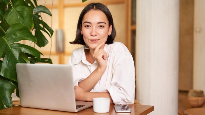 A woman in a white shirt sitting at a café table with a laptop, phone and coffee cup, resting her chin on her hand and smiling.