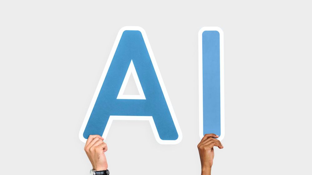 Two raised hands holding up large blue cut-out letters spelling AI against a plain grey wall.