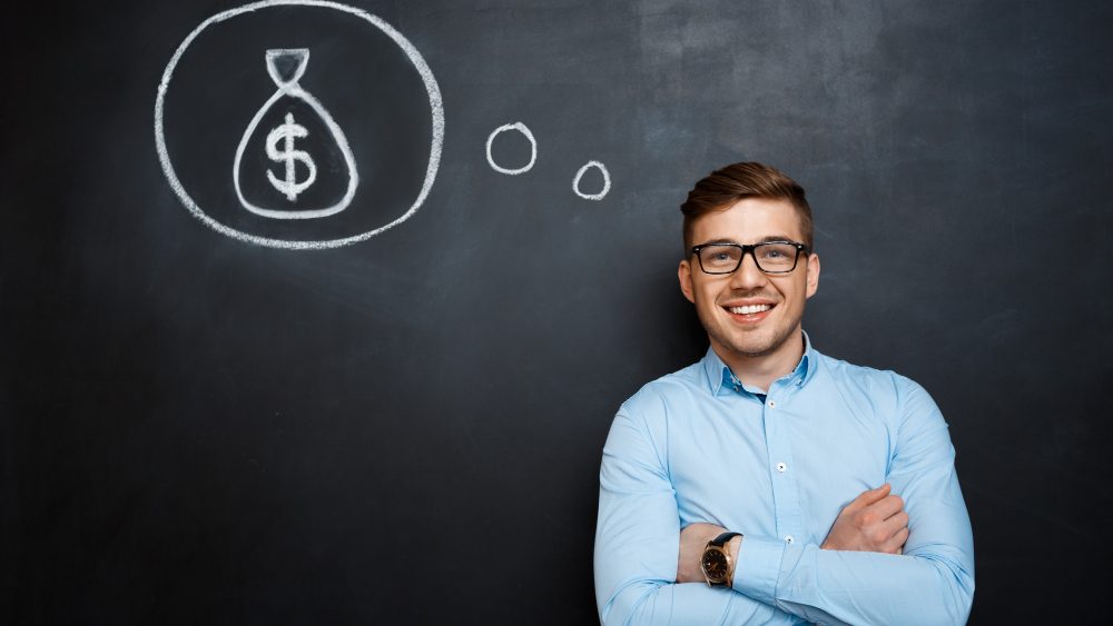 A smiling man in glasses and a blue shirt with arms folded, beside a chalk thought bubble of a money bag on a blackboard.