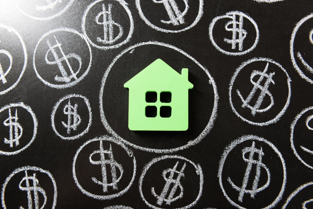 A small green model house on a chalkboard, circled by hand-drawn dollar signs.