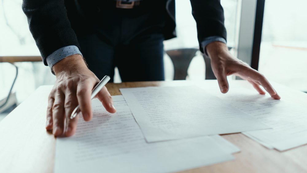 A man in a dark jacket leaning over a table with a pen, reviewing printed pages spread out in front of him.
