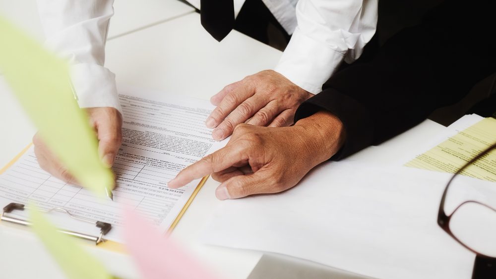 One person pointing to a line on a form clipped to a clipboard while another holds a pen ready to sign it.