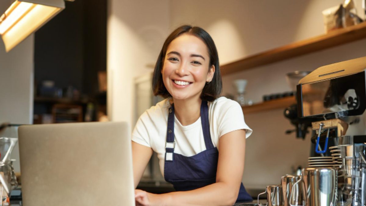 A smiling café owner in an apron leaning on the counter beside an open laptop.