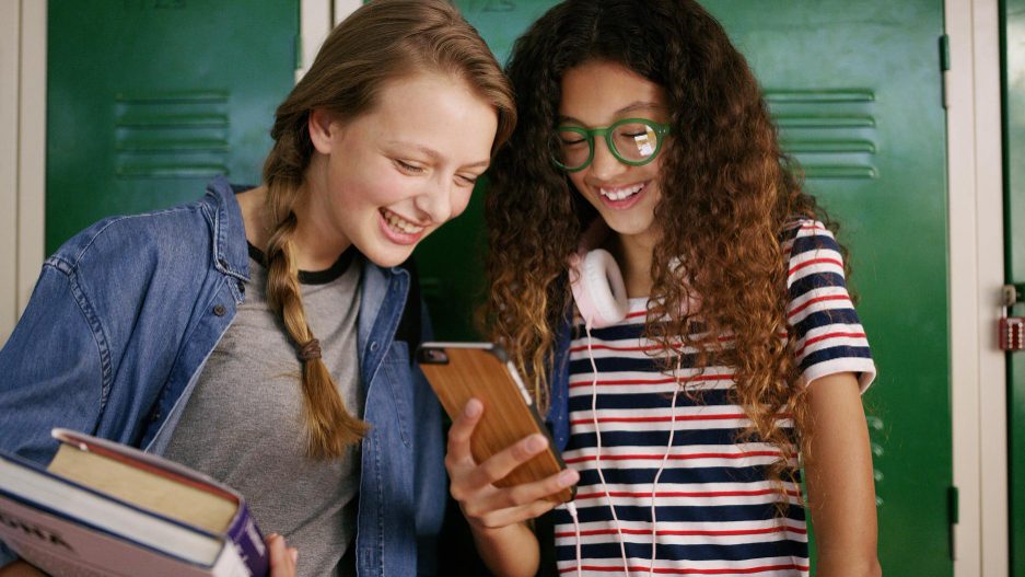 Two teenage girls laughing at a phone together in front of green school lockers, one holding textbooks.