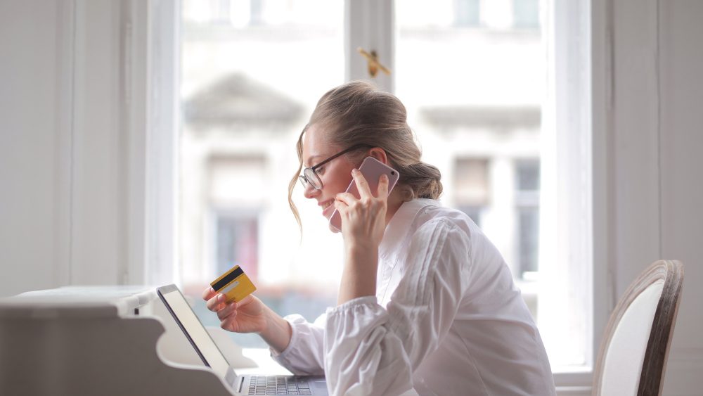 A woman in glasses on the phone at her desk, holding a gold credit card beside an open laptop, in a bright room by a window.