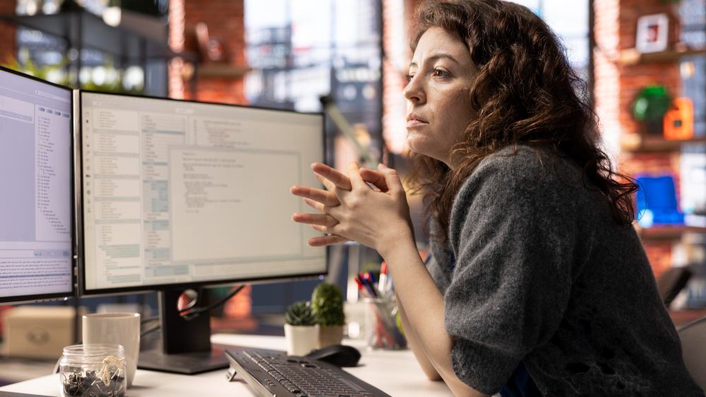 A woman with curly hair leaning on her desk, hands clasped, studying lines of code on two monitors in a brick-walled office.