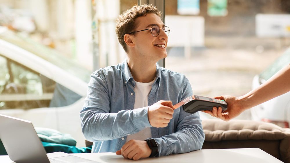 A smiling young man in glasses and a denim shirt tapping a card on a payment terminal held out by a server at a café table.