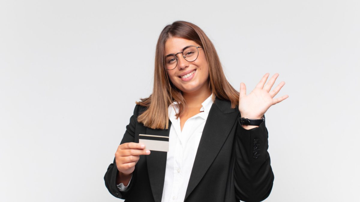A smiling young woman in a blazer holding a credit card and waving.