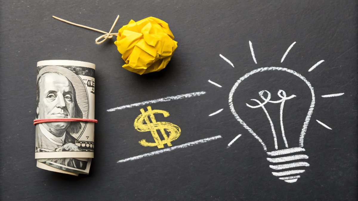 A roll of dollar bills beside a chalk drawing of a light bulb on a blackboard.