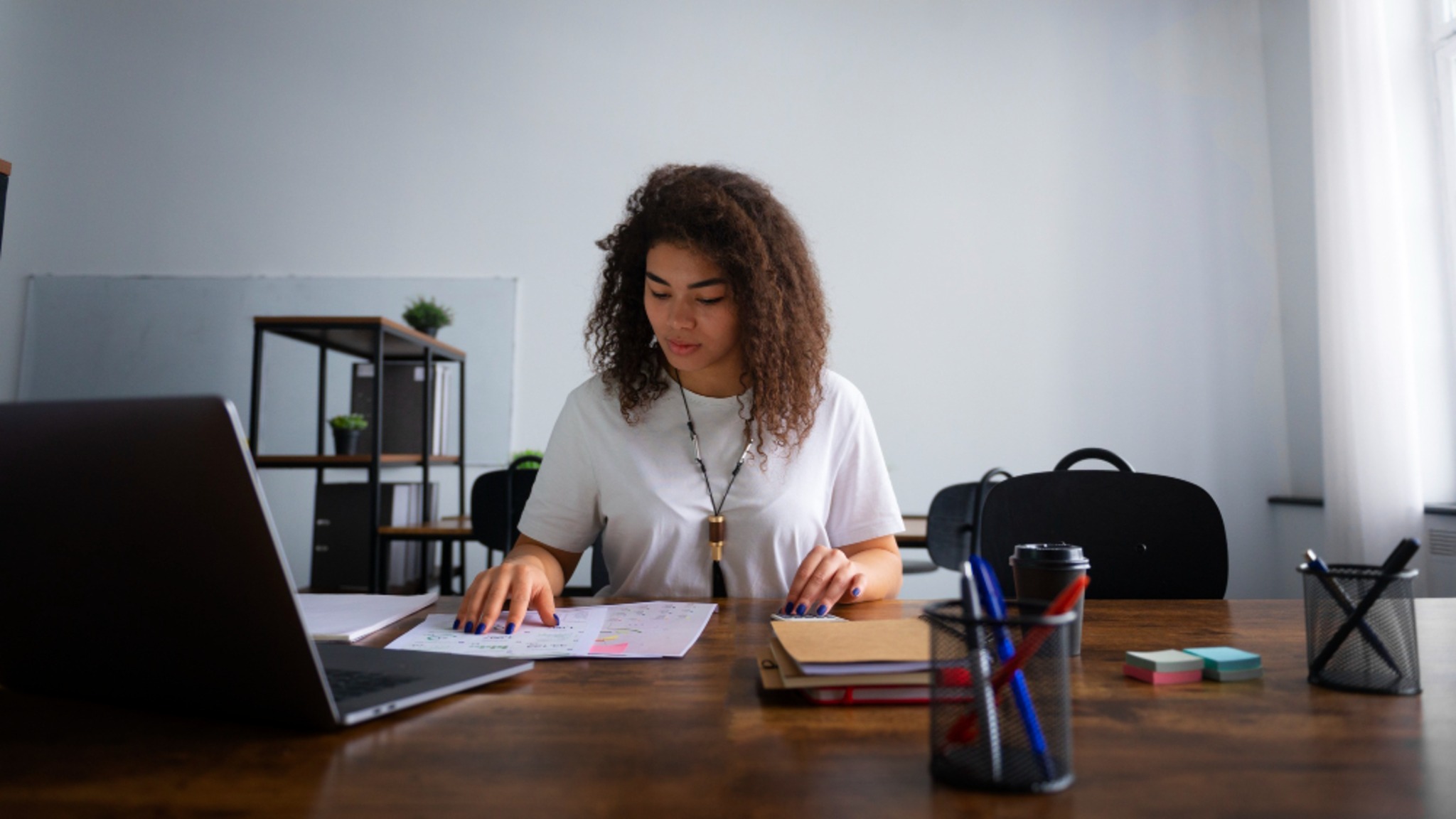 A woman at a desk with a laptop, reviewing printed figures.