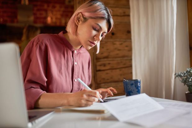 A person at a kitchen table writing on a document beside a laptop and a mug.